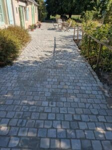 Allée pavée en pierre grise menant à une maison avec volets bleus et verts, ombres d'arbres projetées au soleil.