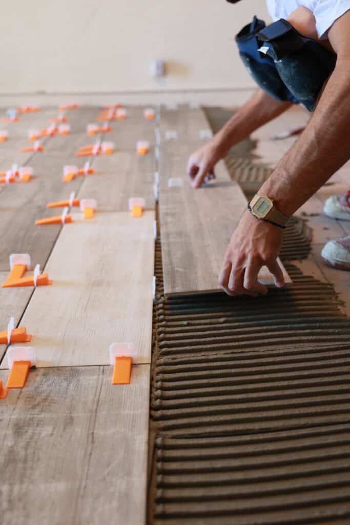 Pose de Carrelage Imitation Bois : Outils et Technique Homme agenouillé posant un carreau imitation bois sur un lit de mortier strié, utilisant des croisillons niveleurs oranges pour le carrelage.