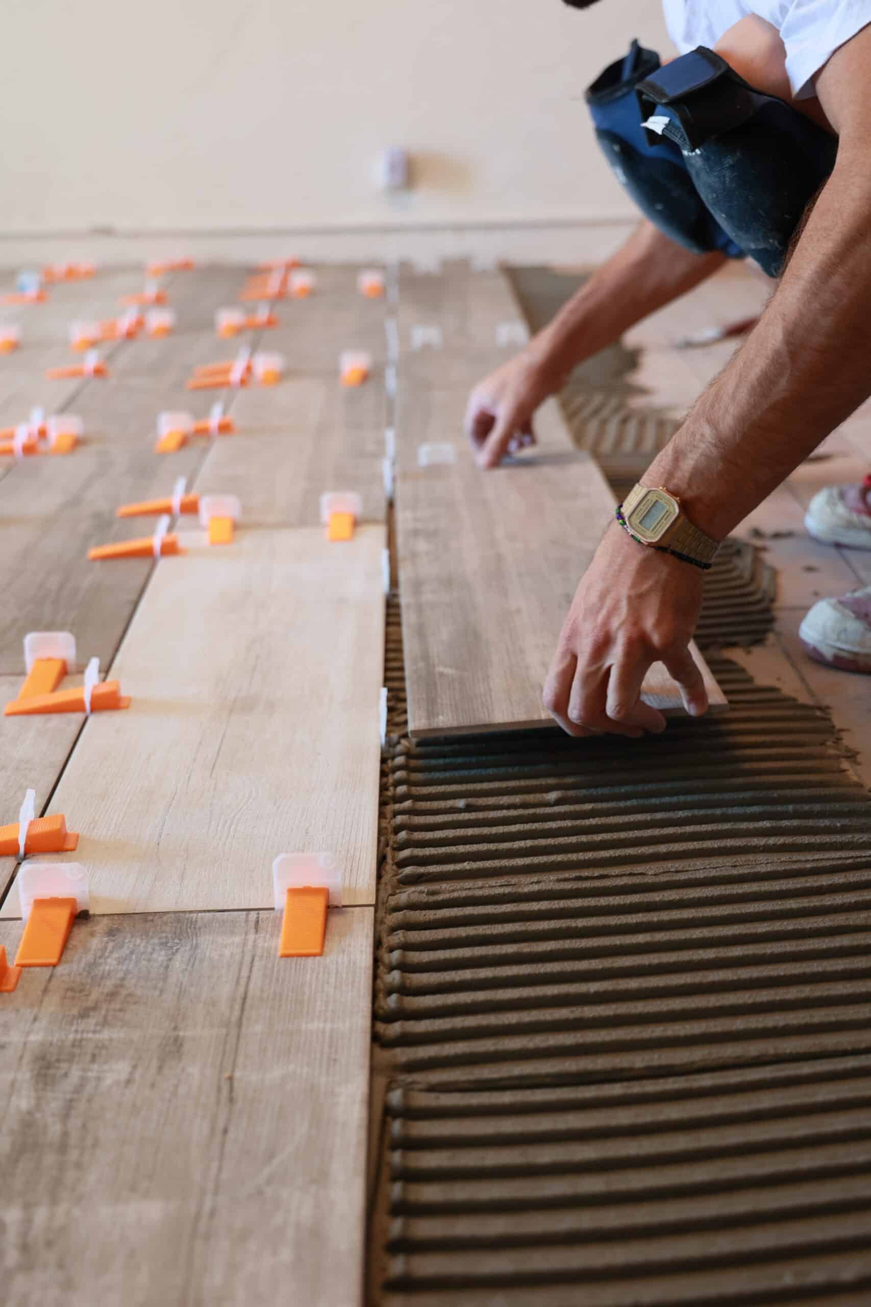 Homme agenouillé posant un carreau imitation bois sur un lit de mortier strié, utilisant des croisillons niveleurs oranges pour le carrelage.