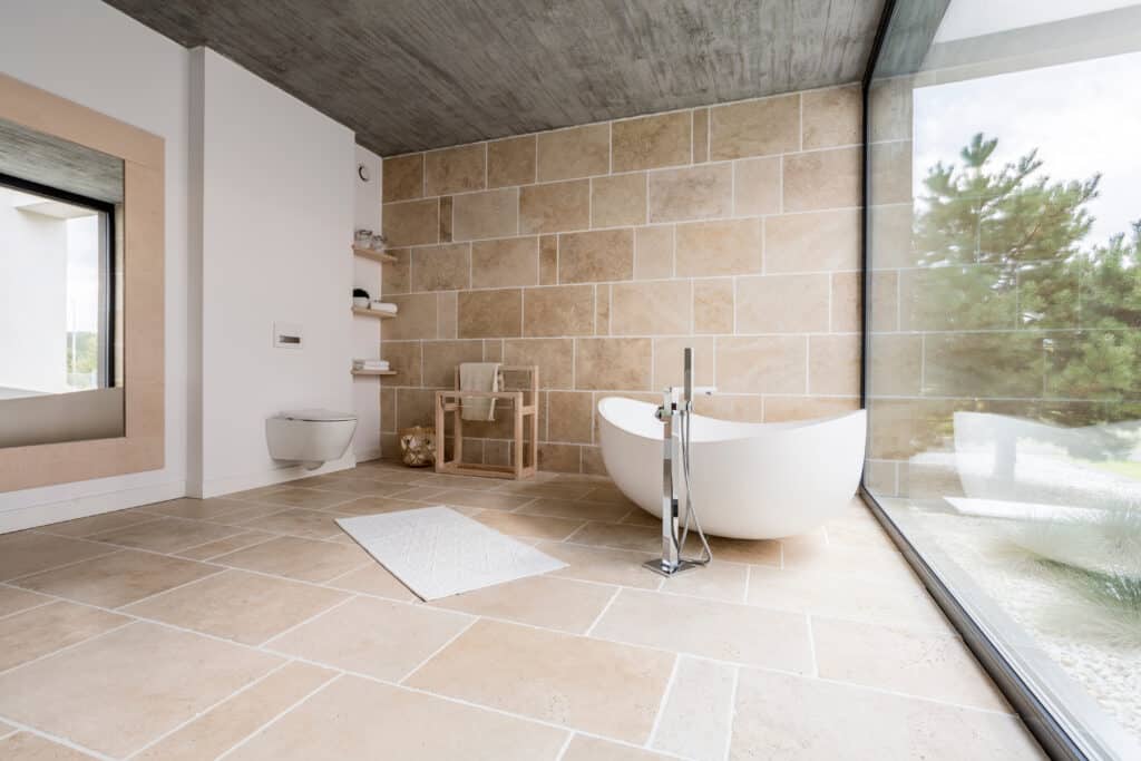 Salle Bain Design Travertin Baignoire Îlot Nature Salle de bain moderne avec baignoire îlot, carrelage en travertin, mur de pierre et grande fenêtre donnant sur la verdure.