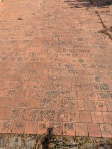 Terrasse en dalles de brique rouge anciennes, tachées de noir, avec mauvaises herbes poussant dans les fissures.