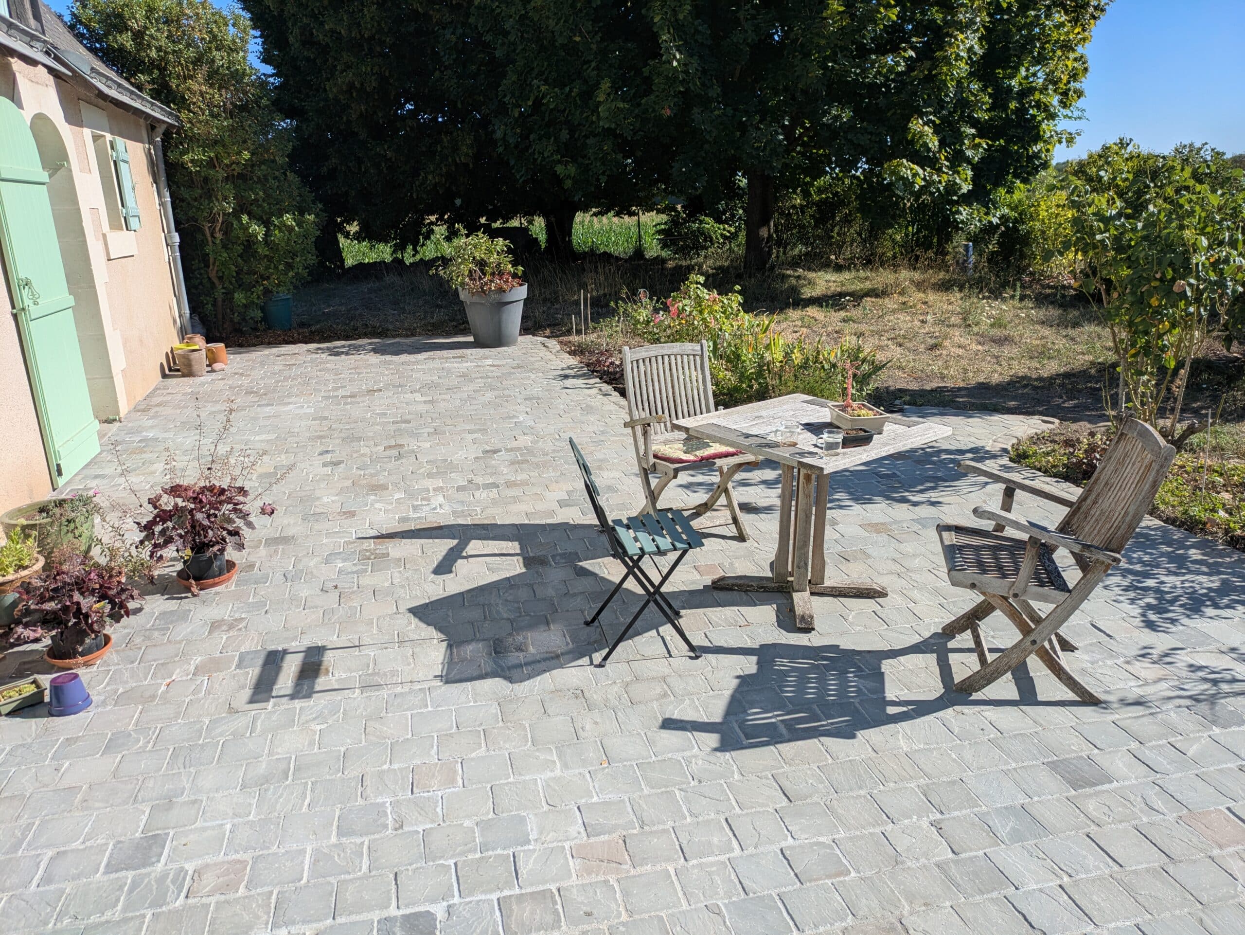 Terrasse pavée de dalles grises devant une maison aux volets vert d'eau, mobilier de jardin en bois sous le soleil.